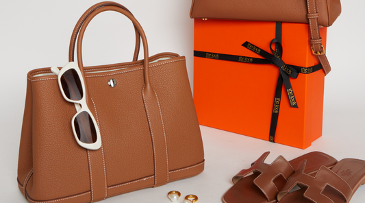 Stylish brown leather bag with sunglasses, gift box, jewelry, and sandals on a white background.