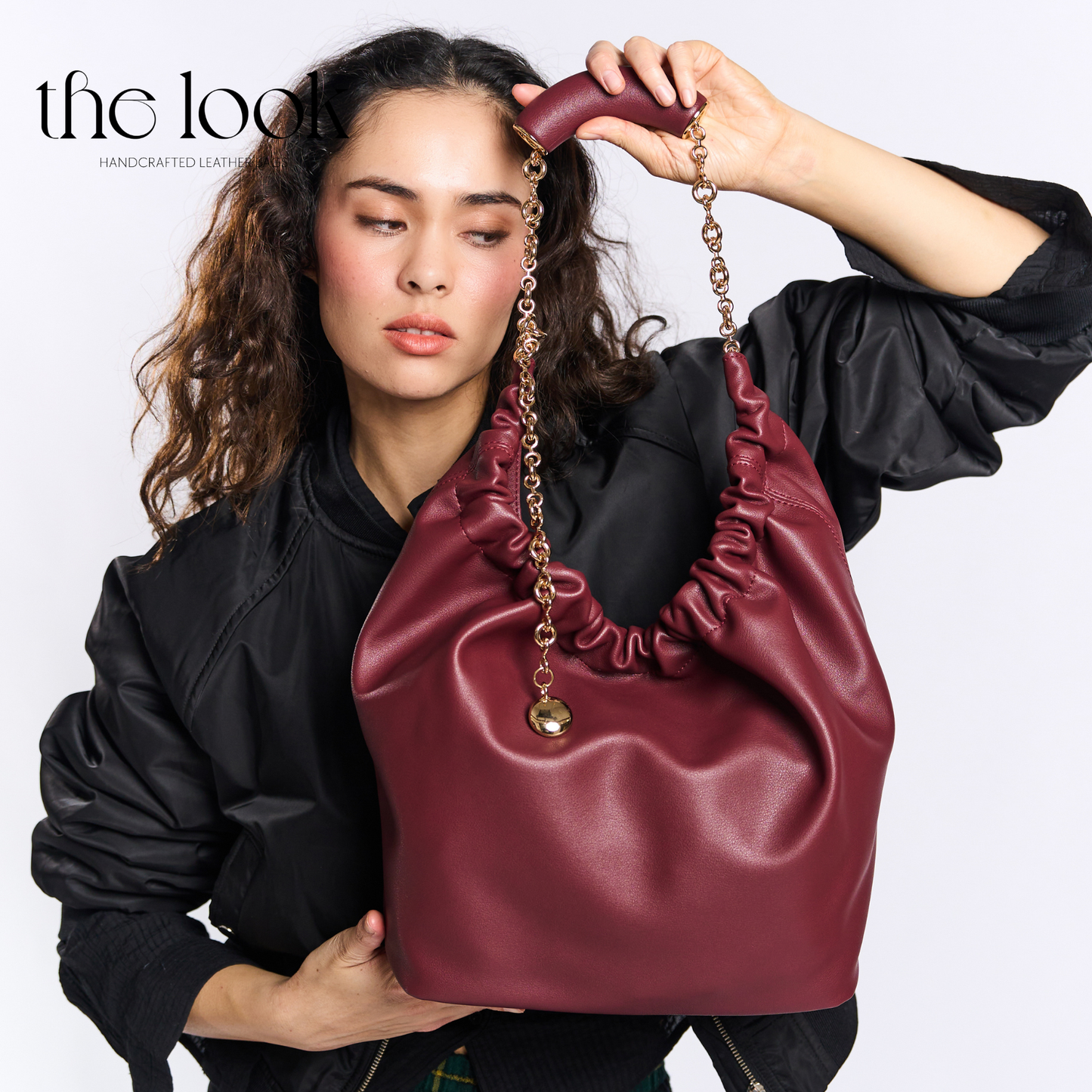 Woman holding a maroon handbag with gold chain detail against a white background. | The Look Officiel