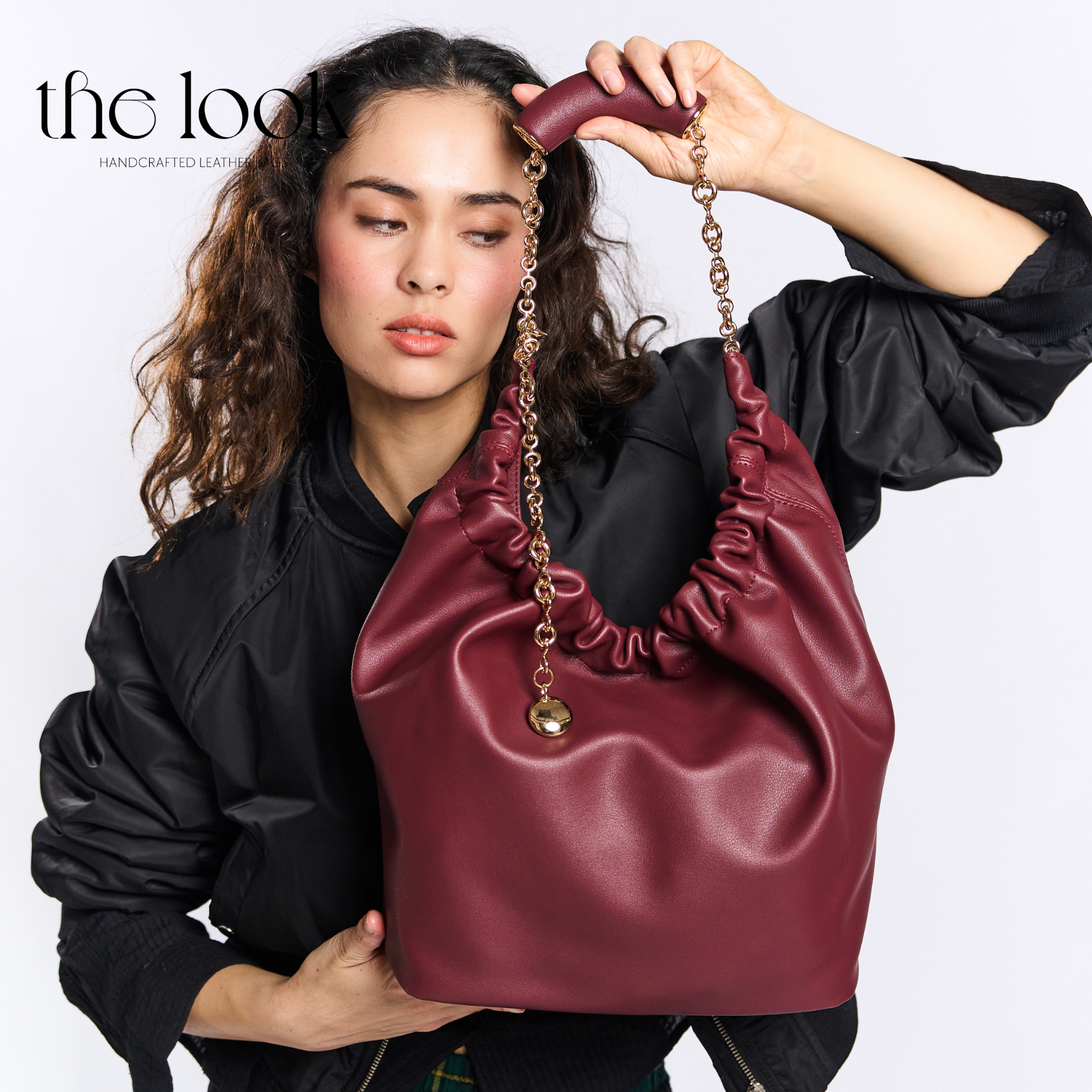 Woman holding a maroon handbag with gold chain detail against a white background. | The Look Officiel