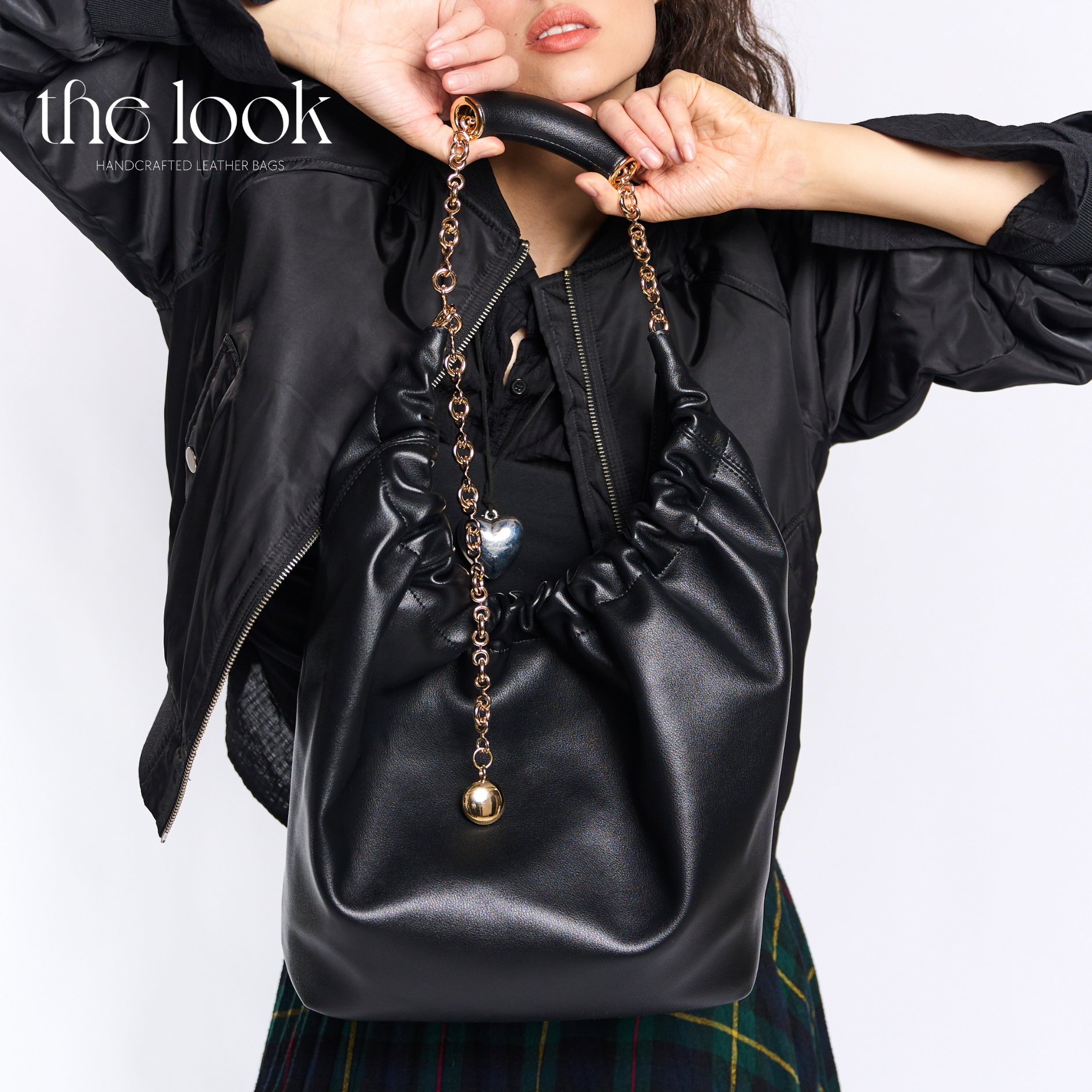 Person holding a black leather bag with decorative chain and heart charm against a white background. | The Look Officiel