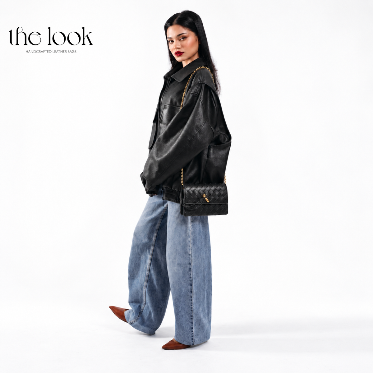 The Loop Wallet on Chain in Black, worn crossbody with a black leather jacket and blue jeans, featuring woven leather and gold-tone hardware.