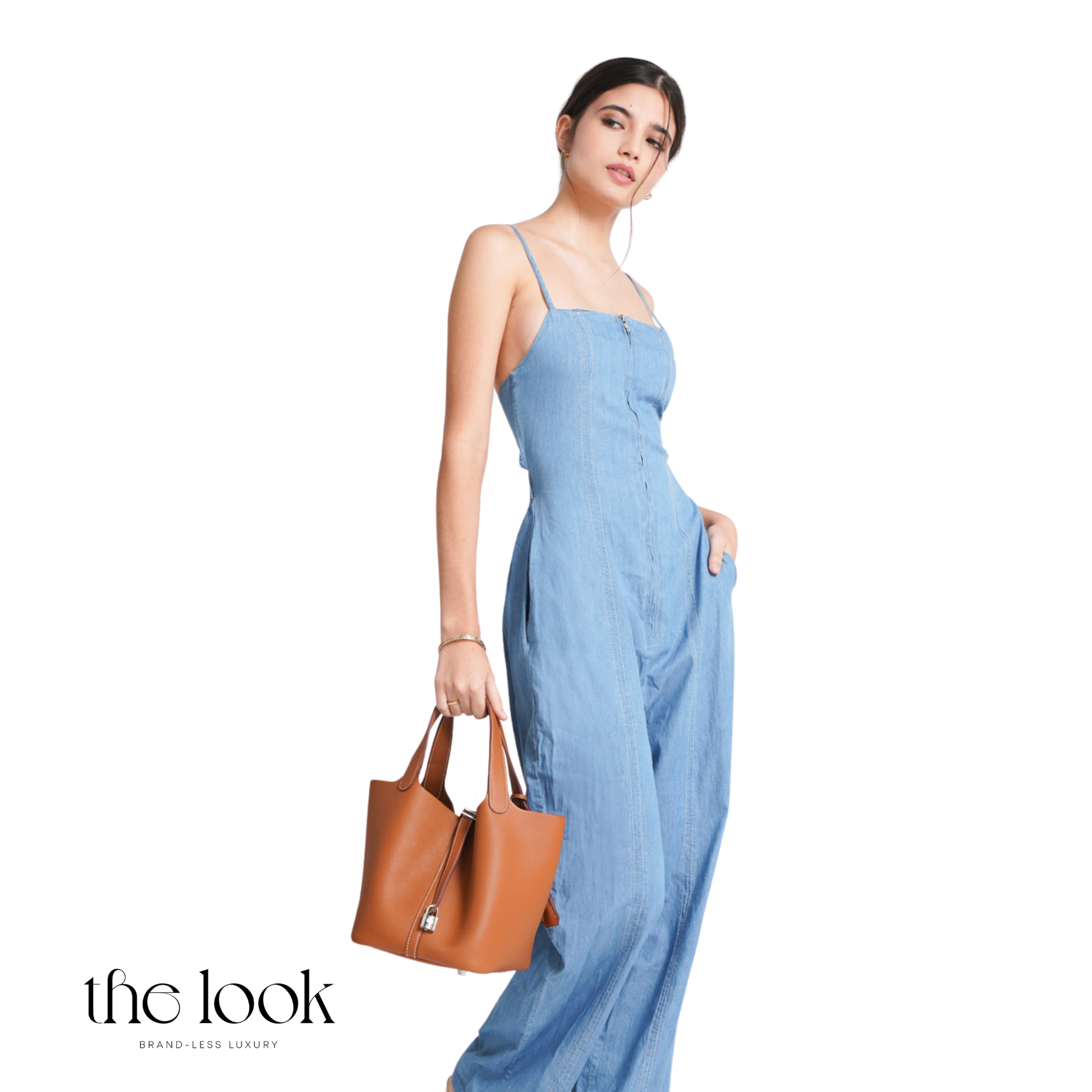 Woman in a blue jumpsuit holding a brown handbag on a white background with 'the look' branding. | The Look Officiel
