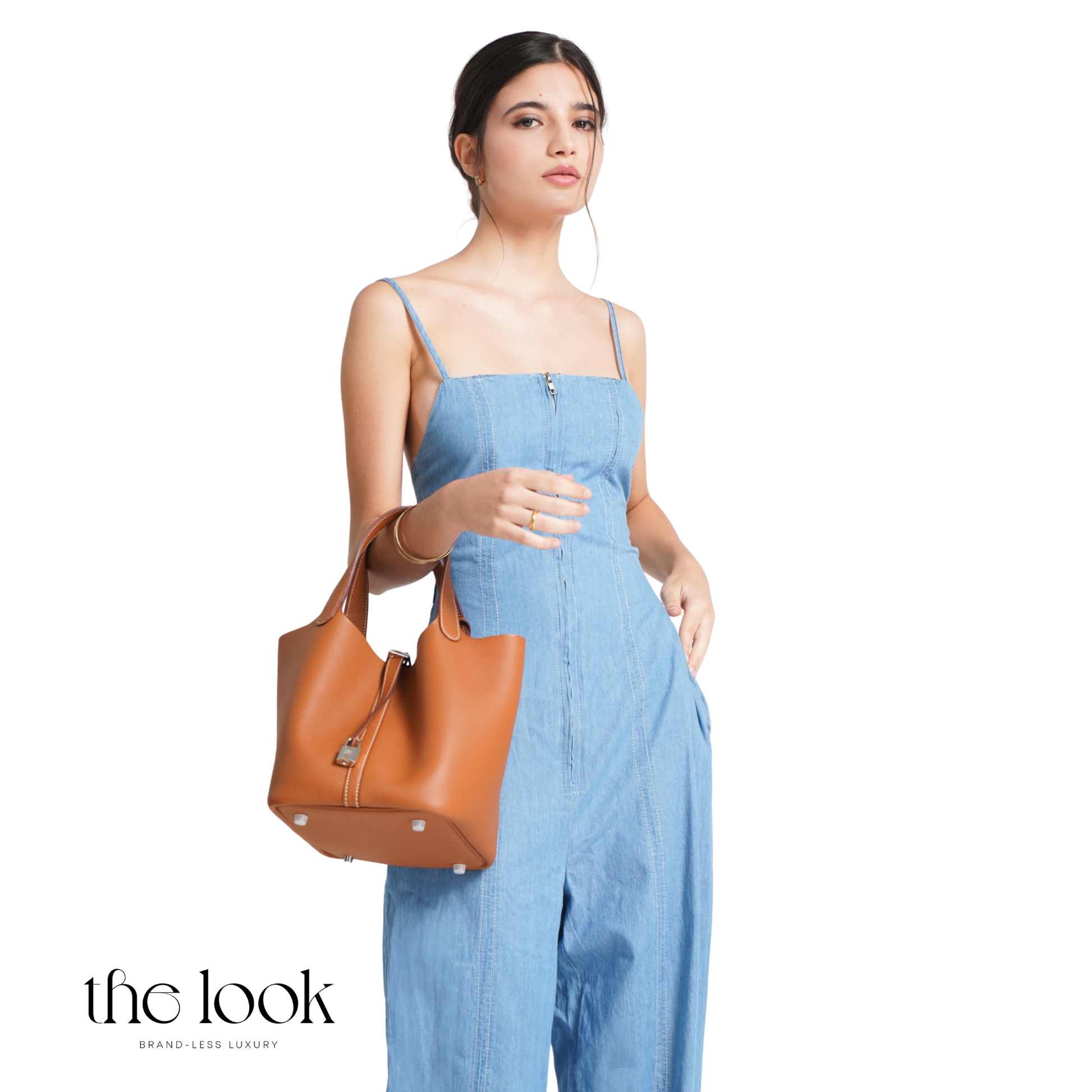 Woman holding a brown handbag wearing a blue jumpsuit on a white background with 'the look' branding. | The Look Officiel