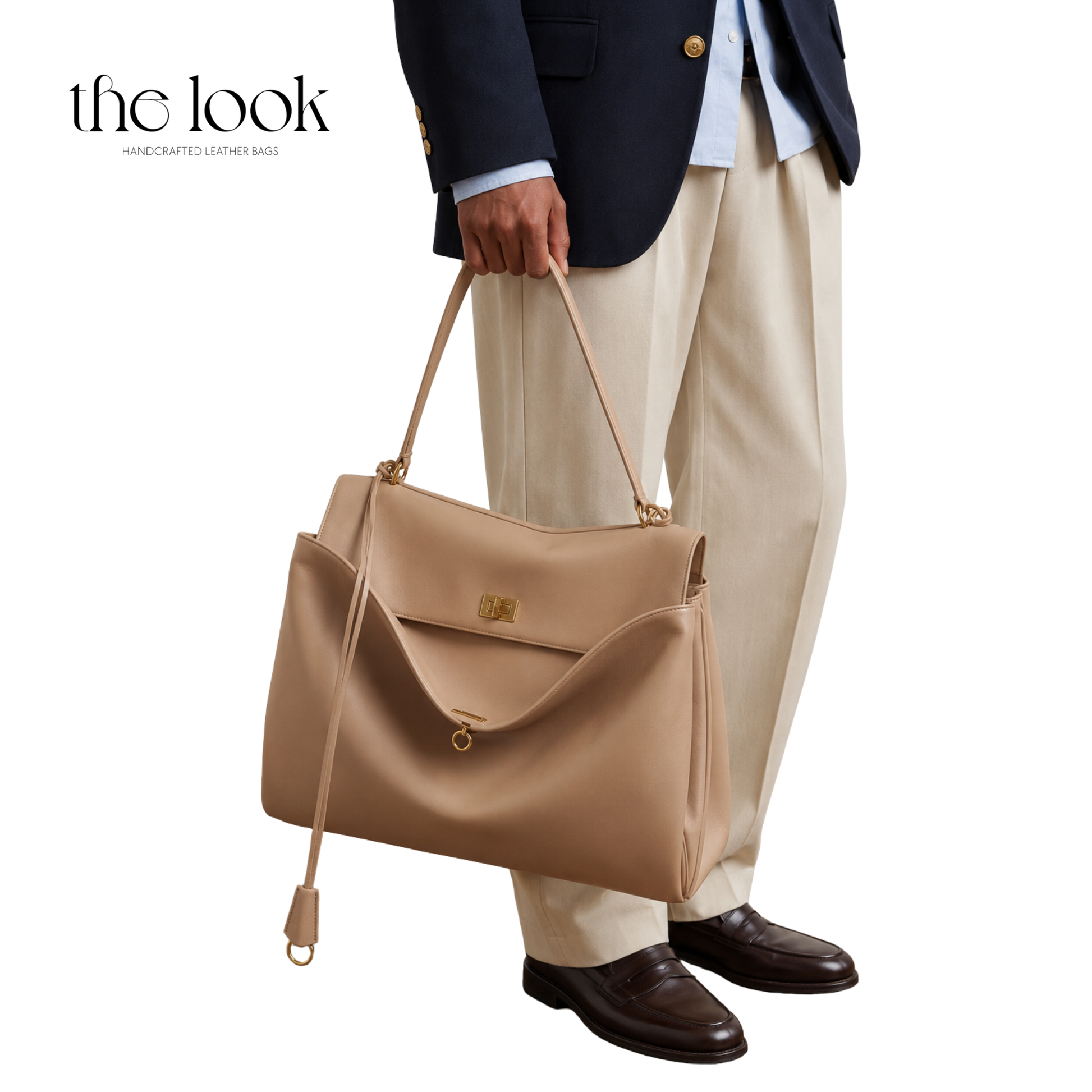 A person wearing a navy blazer, light blue shirt, beige trousers, and dark brown loafers holds a large tan leather tote bag with gold-tone hardware.
