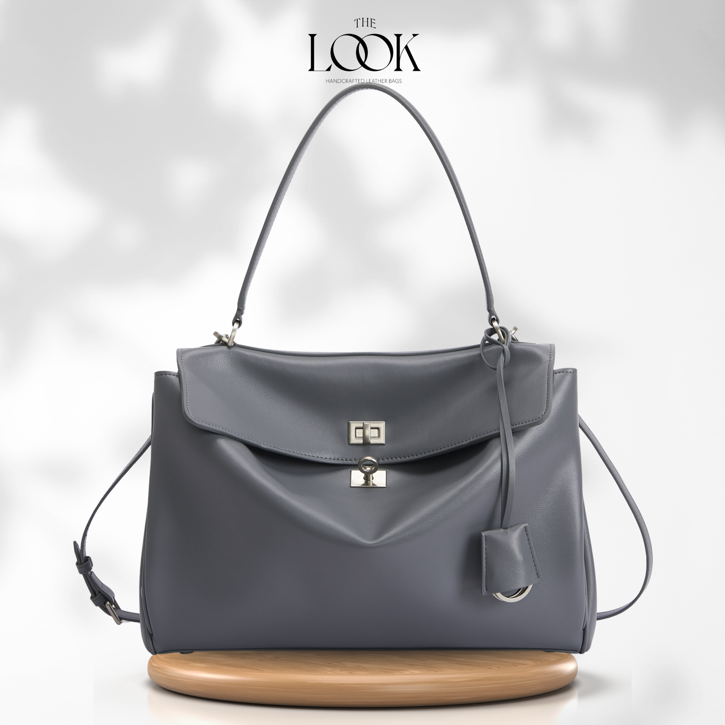 A dark gray leather handbag with a top handle and a small hanging leather tag, displayed on a round wooden stand against a light gray background with soft leaf shadows. The brand name 'THE LOOK' is printed at the top center.